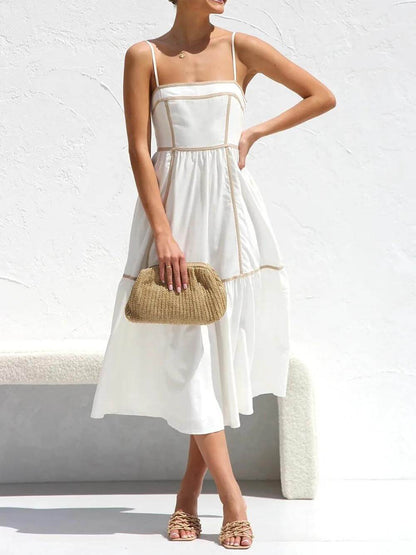 White - A woman wearing a white cotton poplin midi dress with tan contrast piping along the paneled bodice and tiered skirt, holding a woven straw clutch.