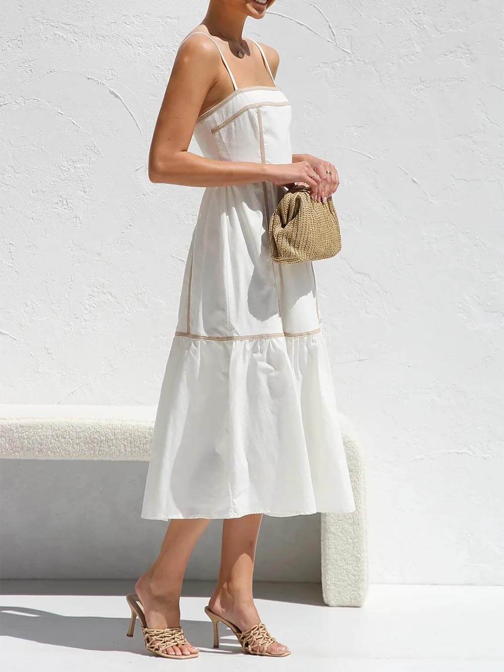 A woman wearing a white cotton poplin midi dress with tan contrast piping along the paneled bodice and tiered skirt, holding a woven straw clutch.