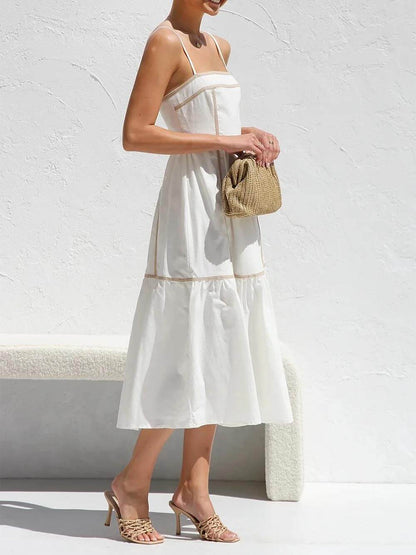 A woman wearing a white cotton poplin midi dress with tan contrast piping along the paneled bodice and tiered skirt, holding a woven straw clutch.