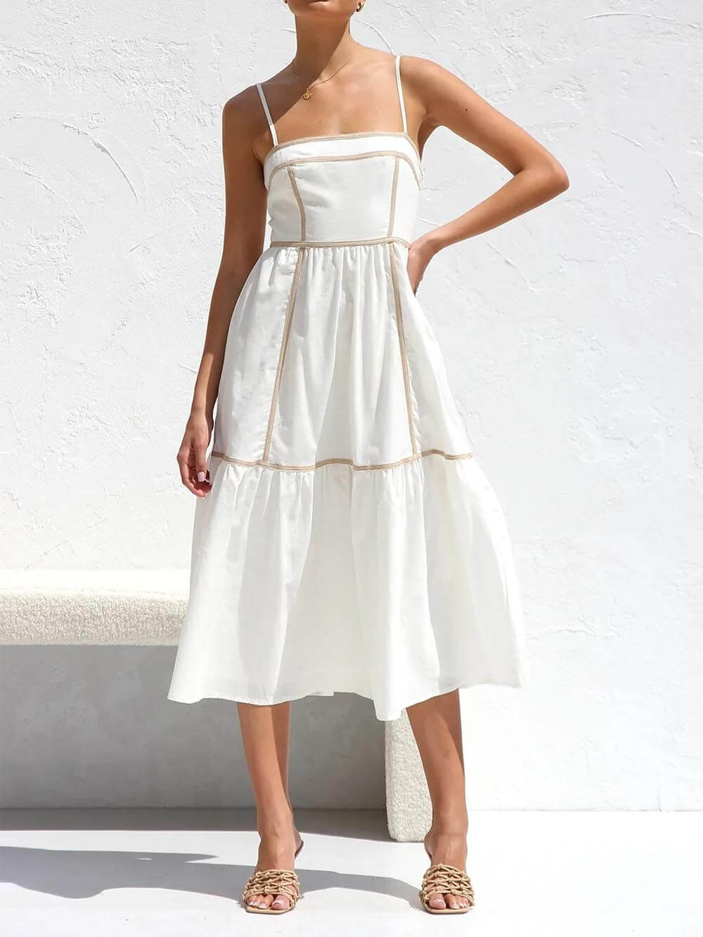A woman wearing a white cotton poplin midi dress with tan contrast piping along the paneled bodice and tiered skirt, holding a woven straw clutch.
