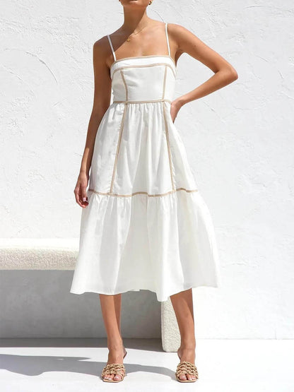 A woman wearing a white cotton poplin midi dress with tan contrast piping along the paneled bodice and tiered skirt, holding a woven straw clutch.