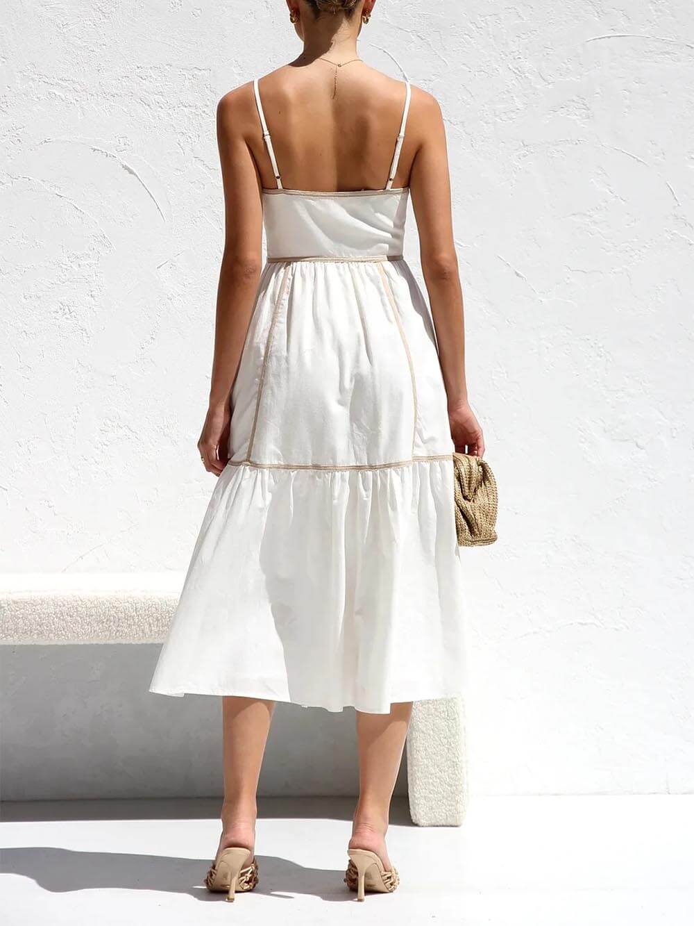 A woman wearing a white cotton poplin midi dress with tan contrast piping along the paneled bodice and tiered skirt, holding a woven straw clutch.