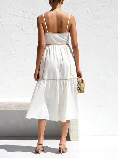 A woman wearing a white cotton poplin midi dress with tan contrast piping along the paneled bodice and tiered skirt, holding a woven straw clutch.