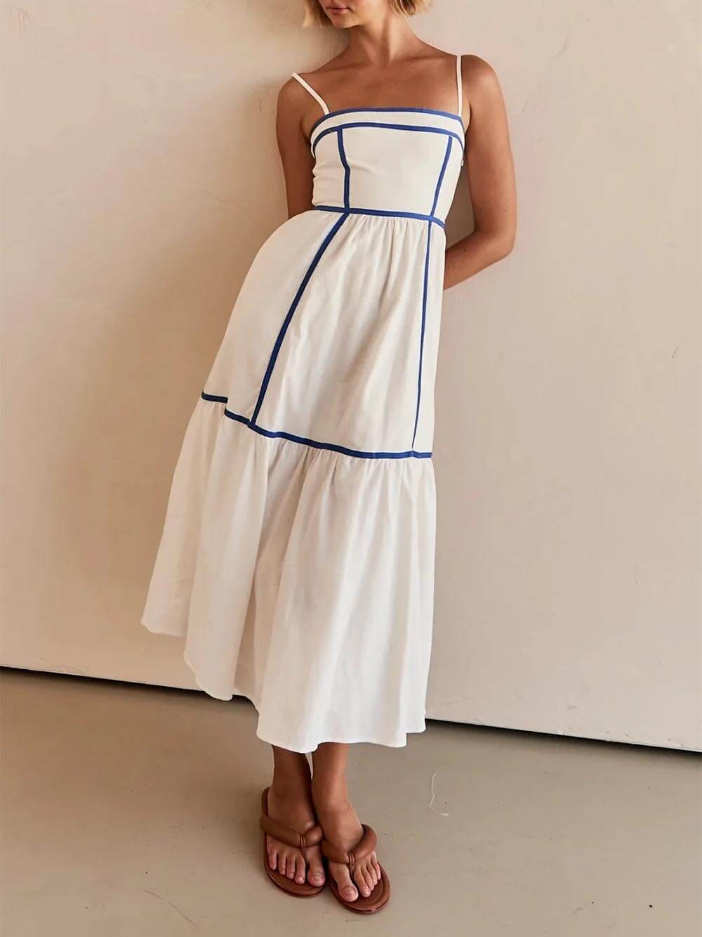 A woman wearing a white cotton poplin midi dress with tan contrast piping along the paneled bodice and tiered skirt, holding a woven straw clutch.