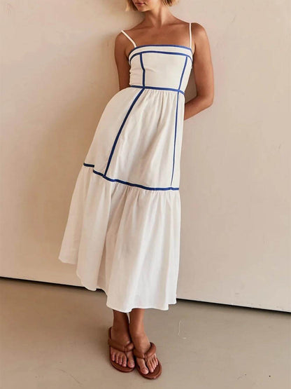 A woman wearing a white cotton poplin midi dress with tan contrast piping along the paneled bodice and tiered skirt, holding a woven straw clutch.