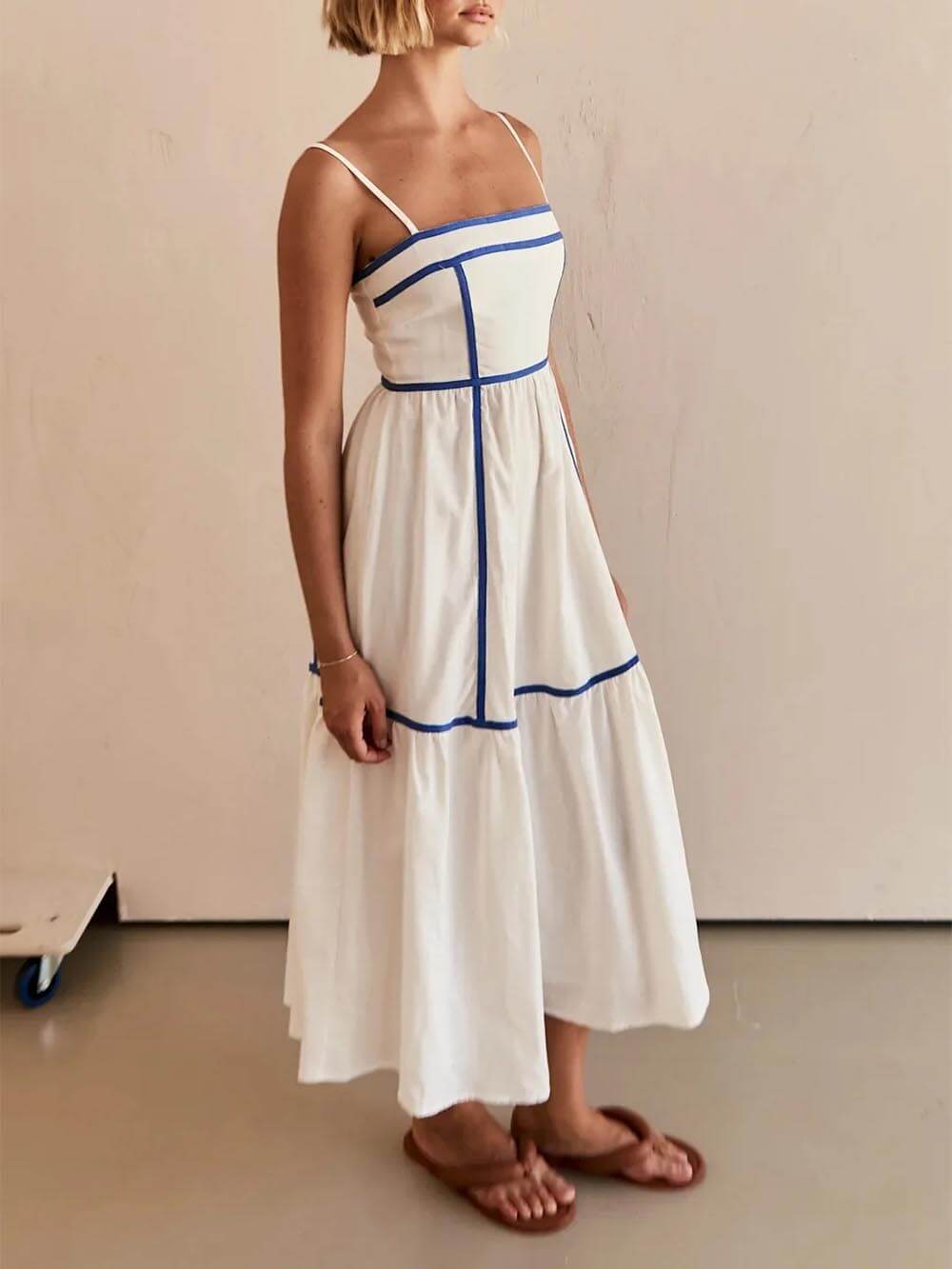 A woman wearing a white cotton poplin midi dress with tan contrast piping along the paneled bodice and tiered skirt, holding a woven straw clutch.