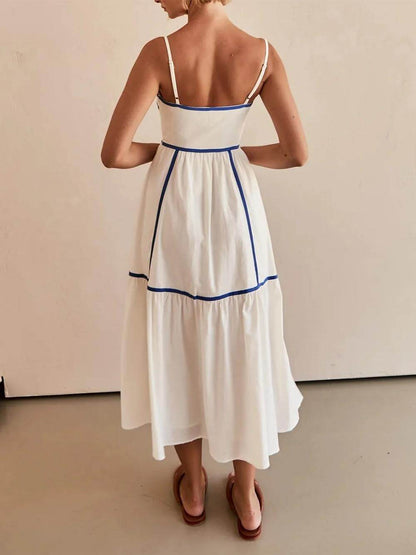 A woman wearing a white cotton poplin midi dress with tan contrast piping along the paneled bodice and tiered skirt, holding a woven straw clutch.