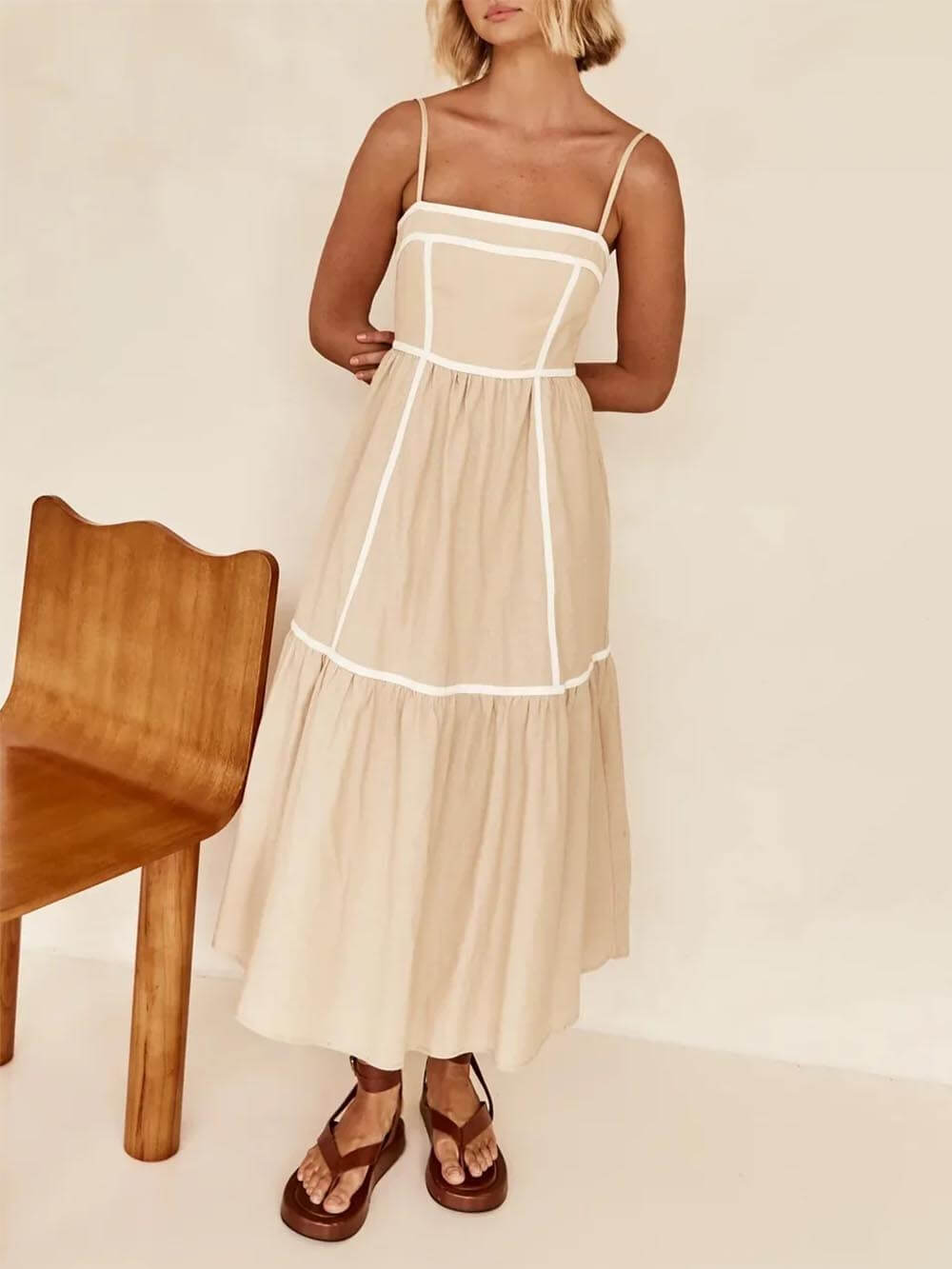 A woman wearing a white cotton poplin midi dress with tan contrast piping along the paneled bodice and tiered skirt, holding a woven straw clutch.