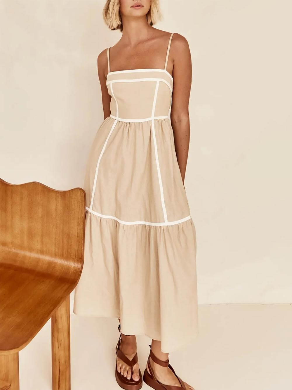 A woman wearing a white cotton poplin midi dress with tan contrast piping along the paneled bodice and tiered skirt, holding a woven straw clutch.