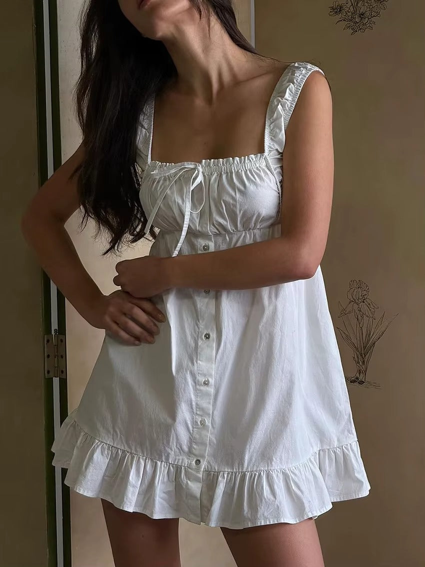 White - A woman wearing a white cotton poplin mini dress featuring a ruched tie-front bust, button-down front, elasticated straps, and a ruffled hemline.