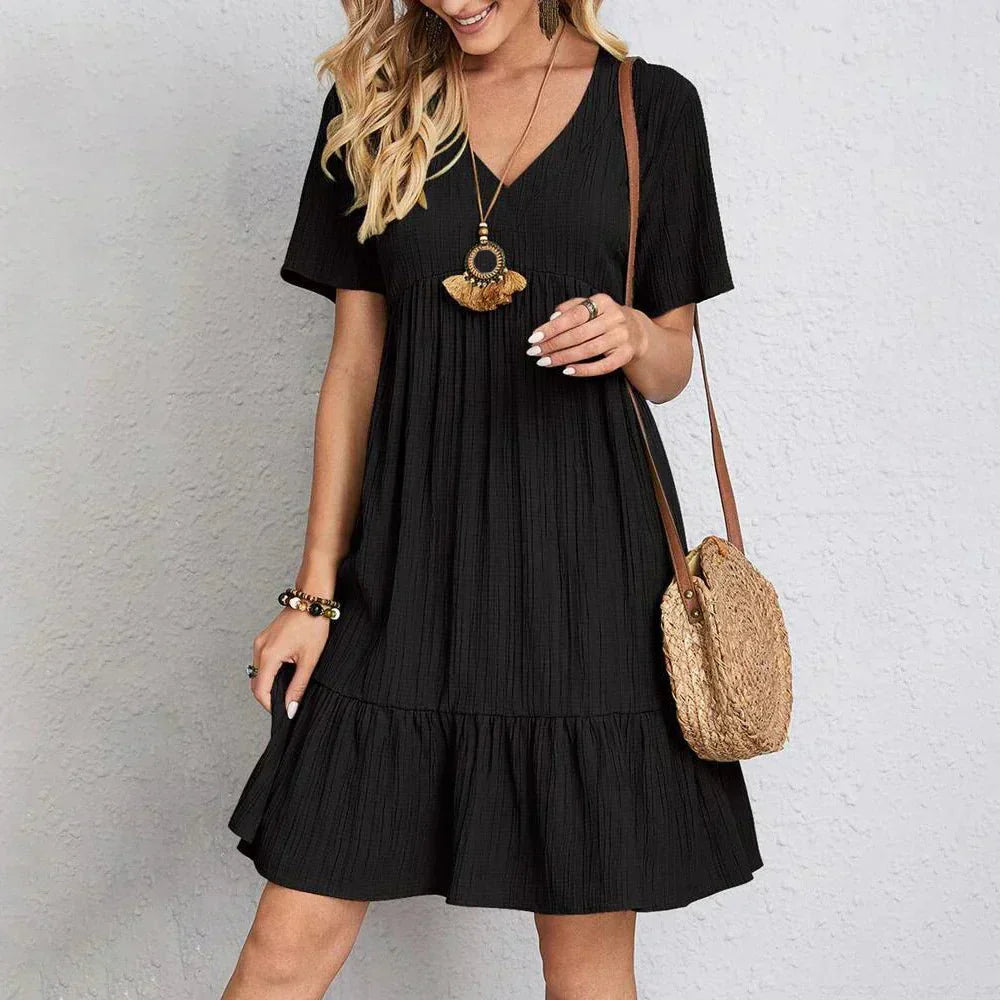 Black - A woman wearing a camel-colored, short-sleeved crinkle gauze mini dress with a V-neckline, empire waist, and tiered ruffled hem, styled with a straw shoulder bag.