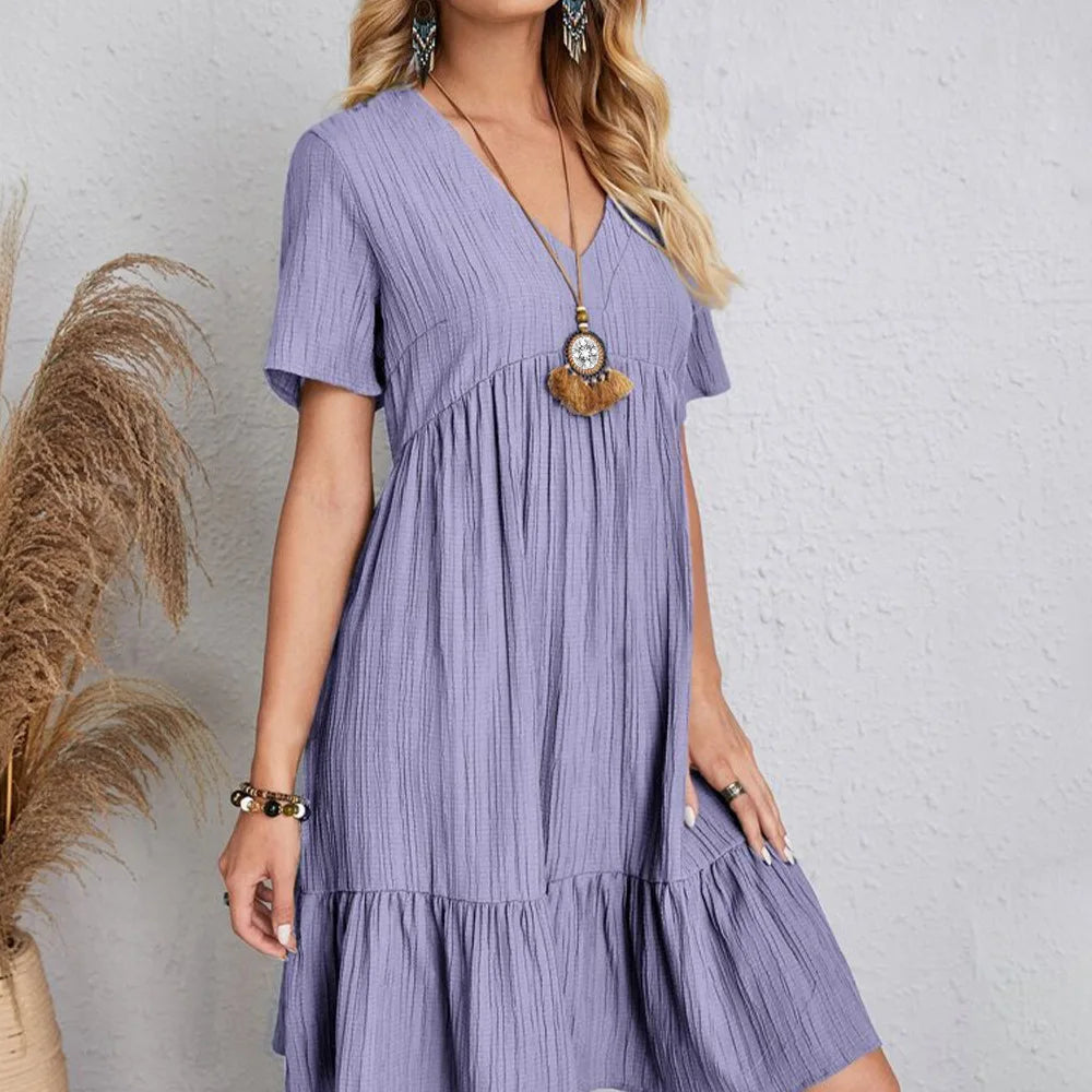 Purple - A woman wearing a camel-colored, short-sleeved crinkle gauze mini dress with a V-neckline, empire waist, and tiered ruffled hem, styled with a straw shoulder bag.