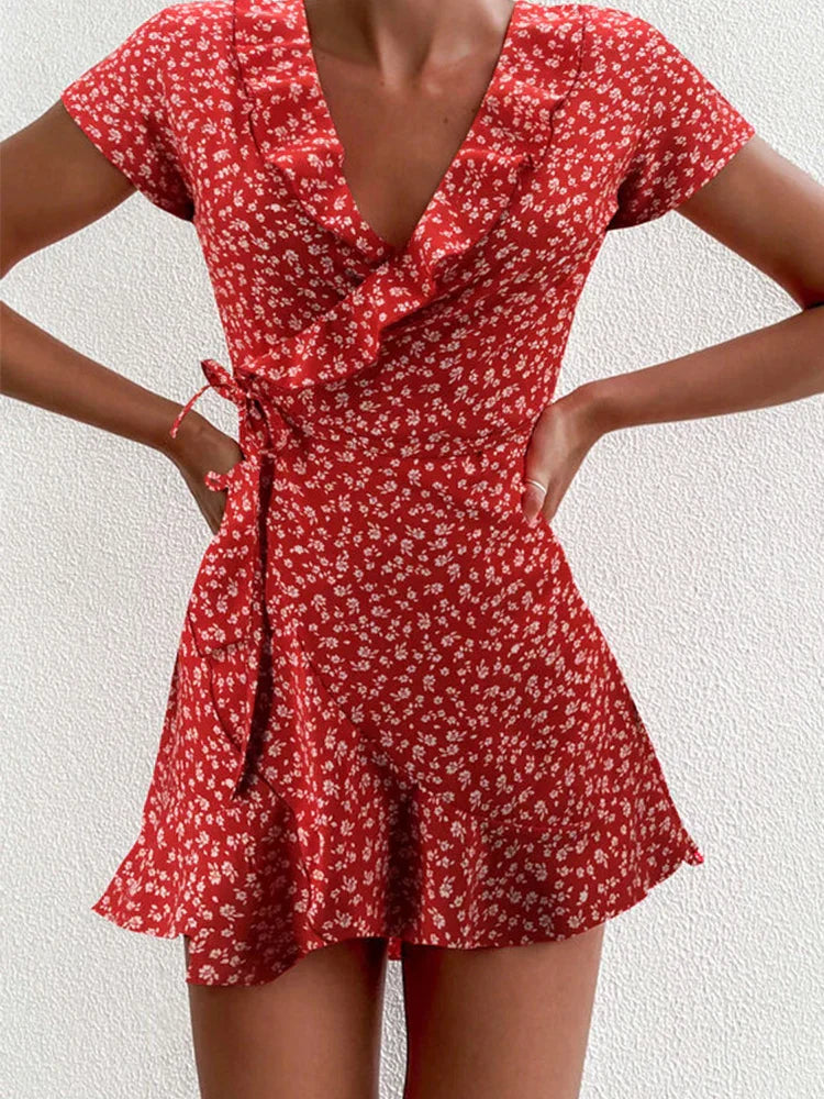 Red - A close-up of a person wearing a red wrap mini dress with a white ditsy floral print, featuring a ruffled V-neckline, short sleeves, and a tie-waist closure.