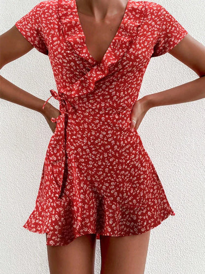 Red - A close-up of a person wearing a red wrap mini dress with a white ditsy floral print, featuring a ruffled V-neckline, short sleeves, and a tie-waist closure.