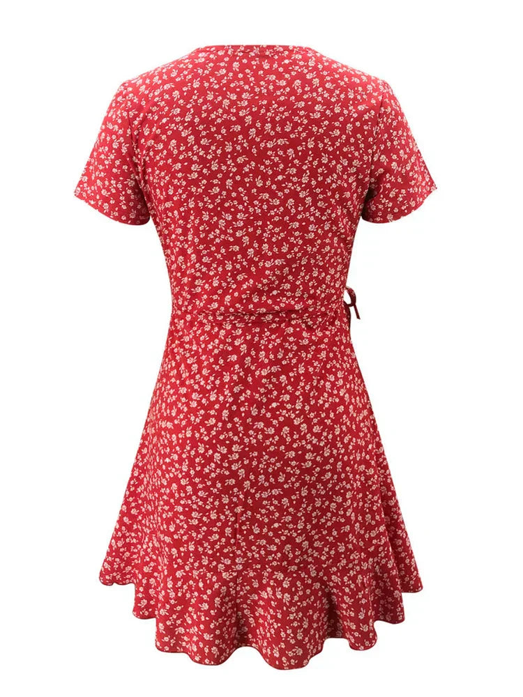 Red - A close-up of a person wearing a red wrap mini dress with a white ditsy floral print, featuring a ruffled V-neckline, short sleeves, and a tie-waist closure.