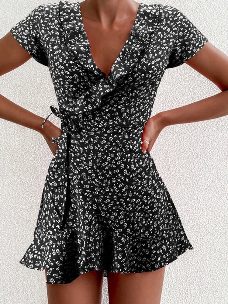 Black - A close-up of a person wearing a red wrap mini dress with a white ditsy floral print, featuring a ruffled V-neckline, short sleeves, and a tie-waist closure.