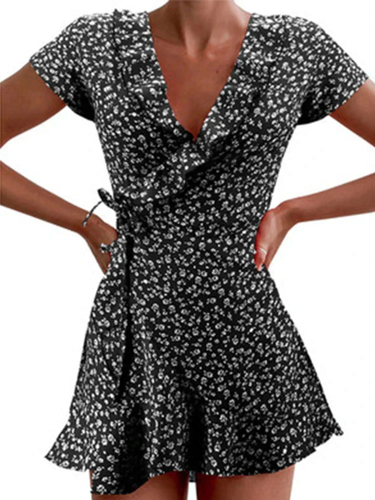 Black - A close-up of a person wearing a red wrap mini dress with a white ditsy floral print, featuring a ruffled V-neckline, short sleeves, and a tie-waist closure.
