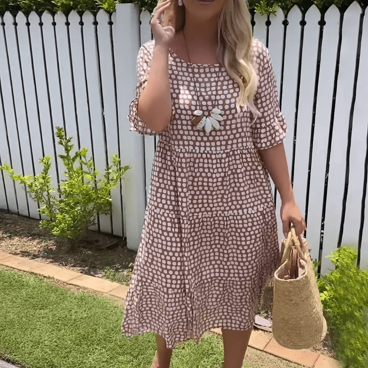 Brown - A woman wearing a taupe and white dot print midi dress with a scoop neck, ruffled half sleeves, and a tiered skirt, holding a woven straw bag.