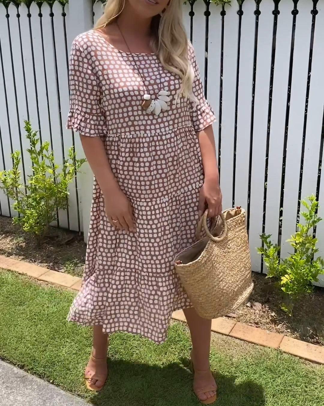 Red - A woman wearing a brown and white dot print tiered midi dress with ruffle-cuffed elbow sleeves, standing outdoors near a white picket fence.