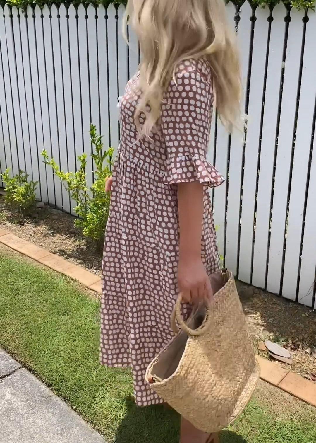 Red - A woman wearing a brown and white dot print tiered midi dress with ruffle-cuffed elbow sleeves, standing outdoors near a white picket fence.