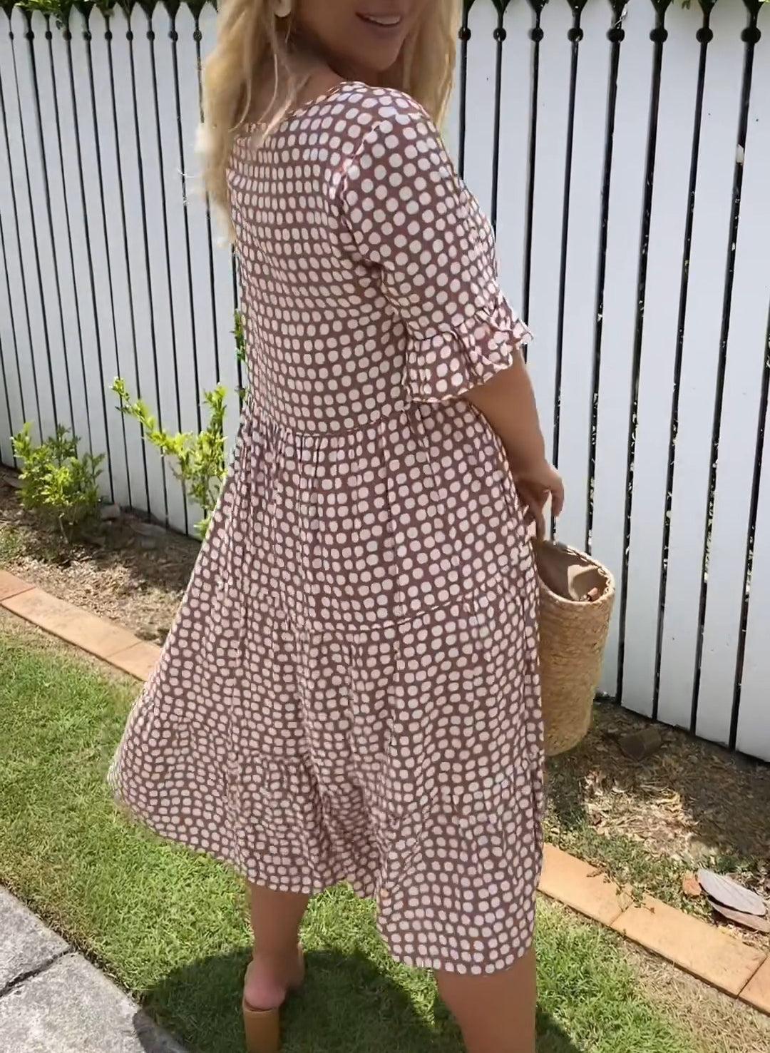 Red - A woman wearing a brown and white dot print tiered midi dress with ruffle-cuffed elbow sleeves, standing outdoors near a white picket fence.