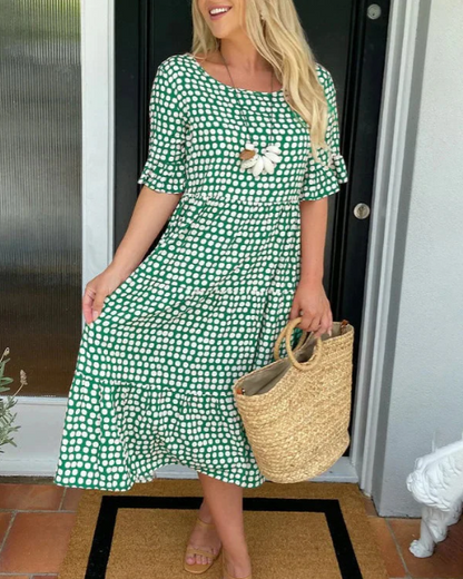 Green - A woman wearing a taupe and white dot print midi dress with a scoop neck, ruffled half sleeves, and a tiered skirt, holding a woven straw bag.