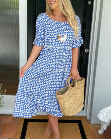 Blue - A woman wearing a taupe and white dot print midi dress with a scoop neck, ruffled half sleeves, and a tiered skirt, holding a woven straw bag.