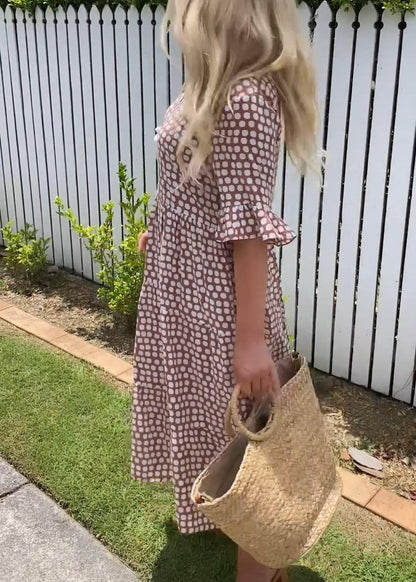 Black - A woman wearing a taupe and white dot print midi dress with a scoop neck, ruffled half sleeves, and a tiered skirt, holding a woven straw bag.