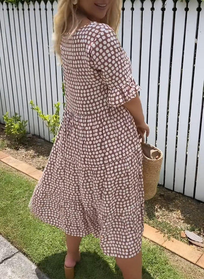 Black - A woman wearing a taupe and white dot print midi dress with a scoop neck, ruffled half sleeves, and a tiered skirt, holding a woven straw bag.