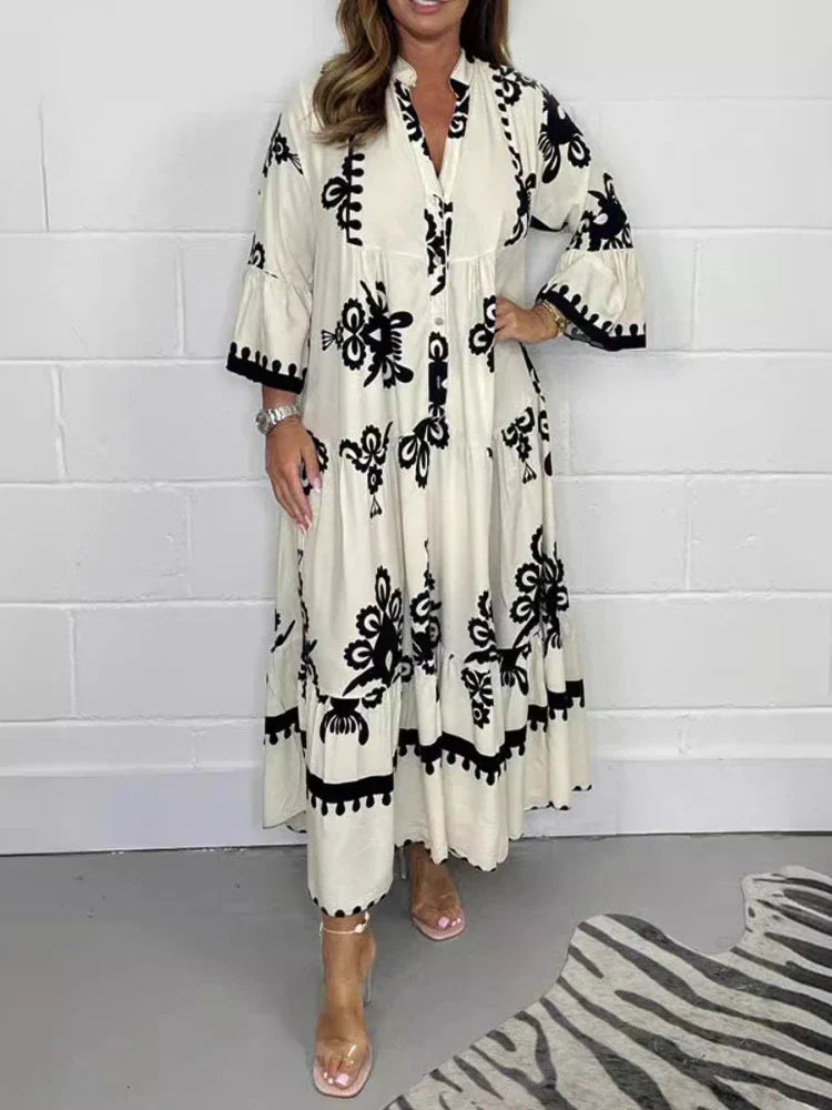 Beige - A woman wearing a black maxi dress with cream-colored contrast embroidery, featuring three-quarter bell sleeves, a split V-neckline, and a tiered skirt with a scalloped hem.