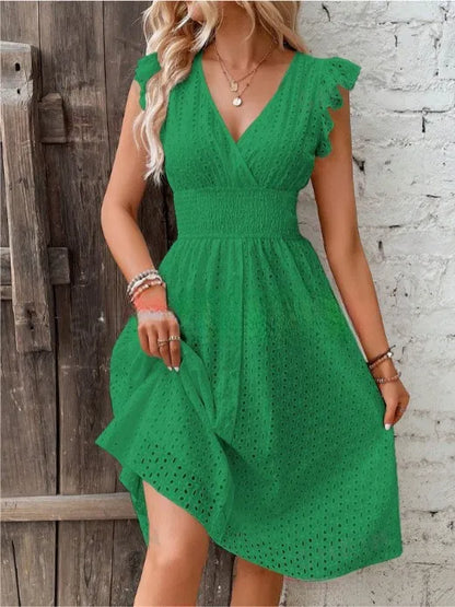 Green - A woman wearing a white eyelet midi dress with flutter sleeves, a surplice V-neckline, and a wide smocked waistband, standing against a rustic wooden door.