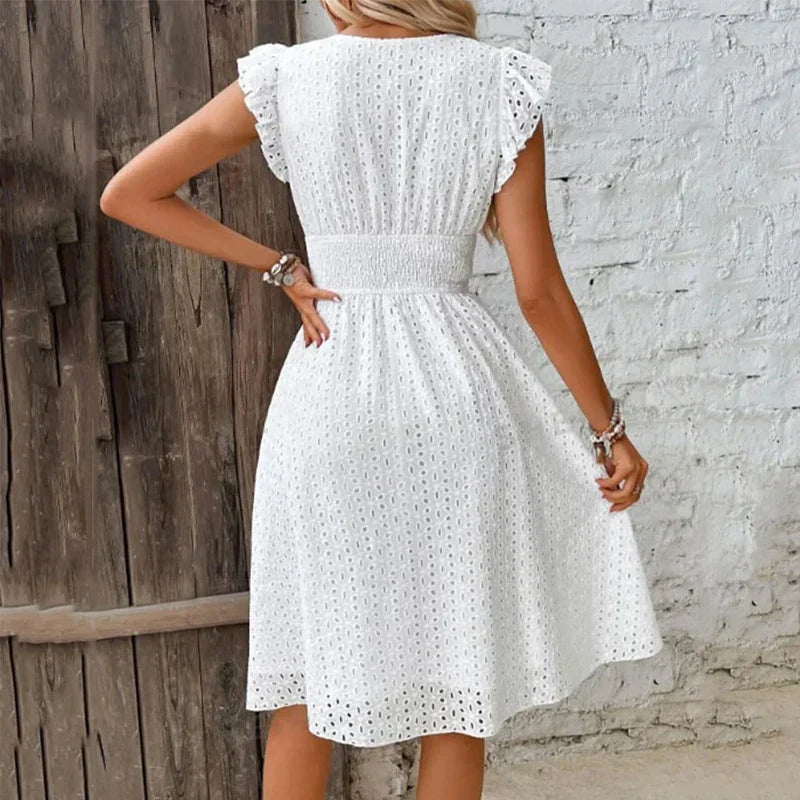 White - A woman wearing a white eyelet midi dress with flutter sleeves, a surplice V-neckline, and a wide smocked waistband, standing against a rustic wooden door.