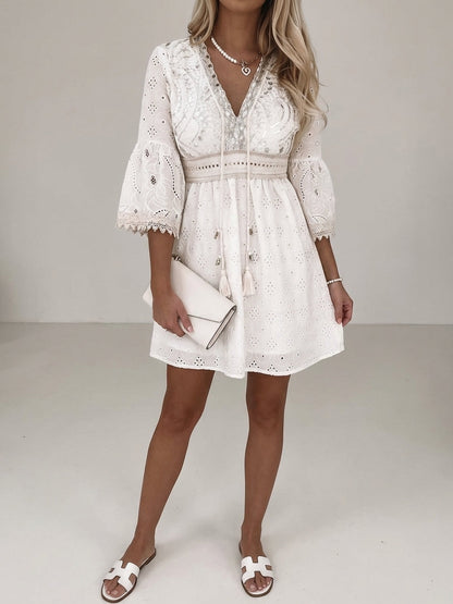 Beige - A woman wearing a cream-colored eyelet lace mini dress with three-quarter bell sleeves, a V-neckline with tassel ties, and a tiered skirt with scalloped lace trim.