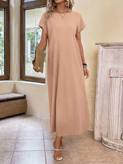 A woman wearing a beige fine-knit maxi dress with cap sleeves and a crew neck, styled with gold jewelry and a woven clutch in a minimalist interior.