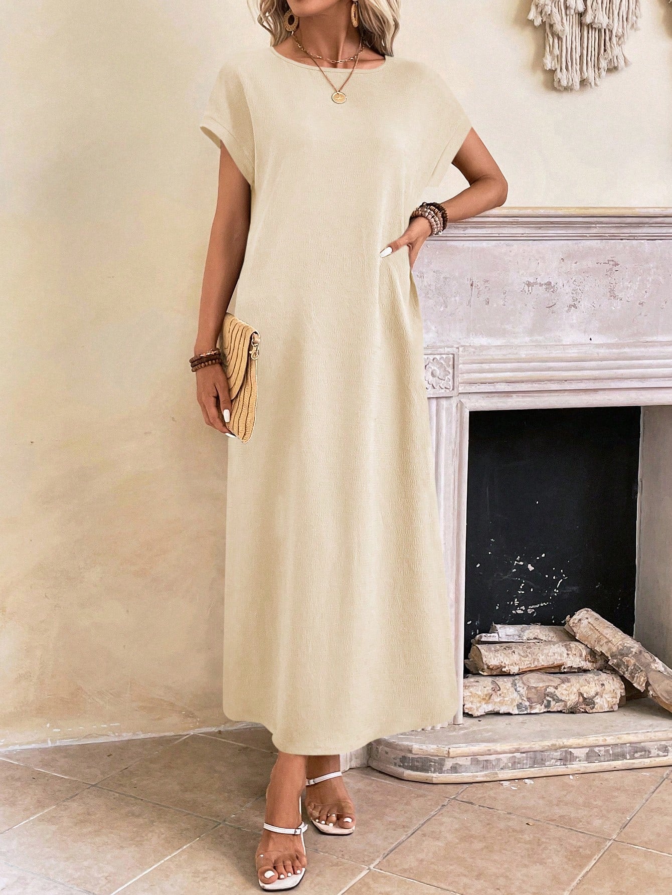Apricot - A woman wearing a beige fine-knit maxi dress with cap sleeves and a crew neck, styled with gold jewelry and a woven clutch in a minimalist interior.