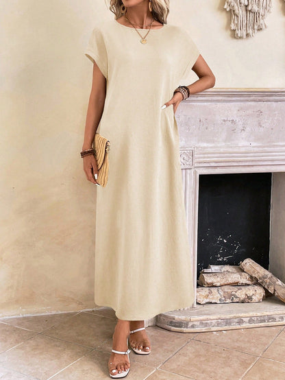 Apricot - A woman wearing a beige fine-knit maxi dress with cap sleeves and a crew neck, styled with gold jewelry and a woven clutch in a minimalist interior.
