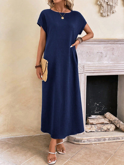 Dark Blue - A woman wearing a beige fine-knit maxi dress with cap sleeves and a crew neck, styled with gold jewelry and a woven clutch in a minimalist interior.