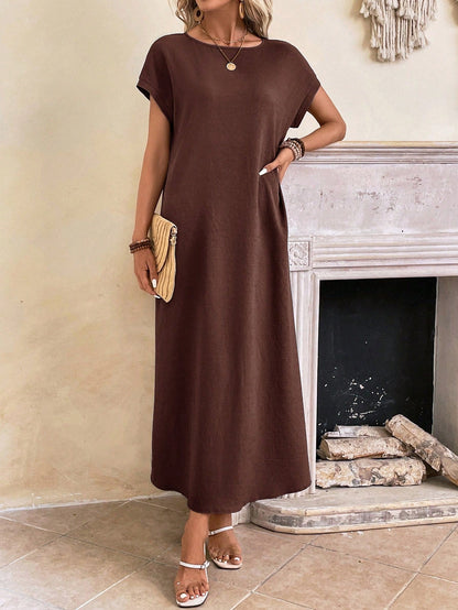 Dark Blue - A woman wearing a beige fine-knit maxi dress with cap sleeves and a crew neck, styled with gold jewelry and a woven clutch in a minimalist interior.