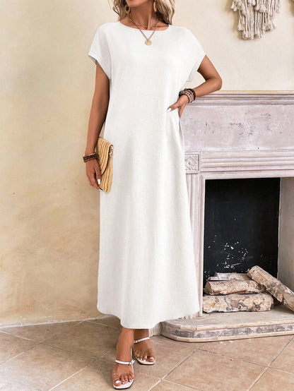 Dark Blue - A woman wearing a beige fine-knit maxi dress with cap sleeves and a crew neck, styled with gold jewelry and a woven clutch in a minimalist interior.