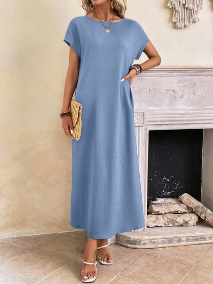 Dark Blue - A woman wearing a beige fine-knit maxi dress with cap sleeves and a crew neck, styled with gold jewelry and a woven clutch in a minimalist interior.