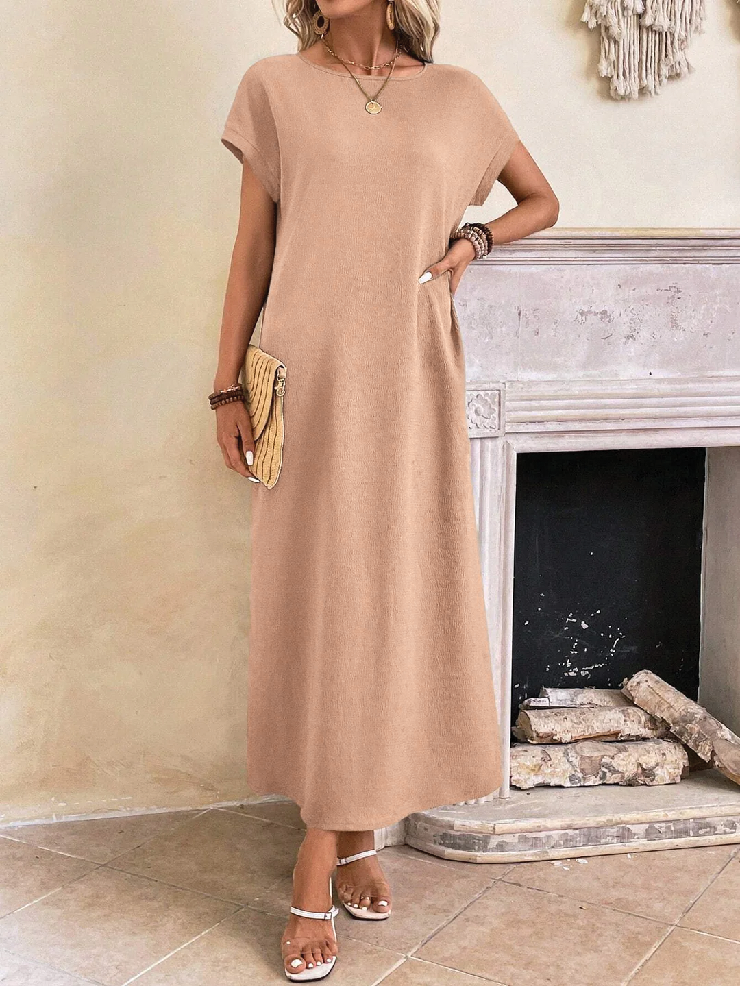 Peachy Pink - A woman wearing a beige fine-knit maxi dress with cap sleeves and a crew neck, styled with gold jewelry and a woven clutch in a minimalist interior.