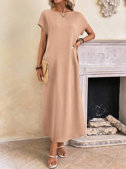 Peachy Pink - A woman wearing a beige fine-knit maxi dress with cap sleeves and a crew neck, styled with gold jewelry and a woven clutch in a minimalist interior.