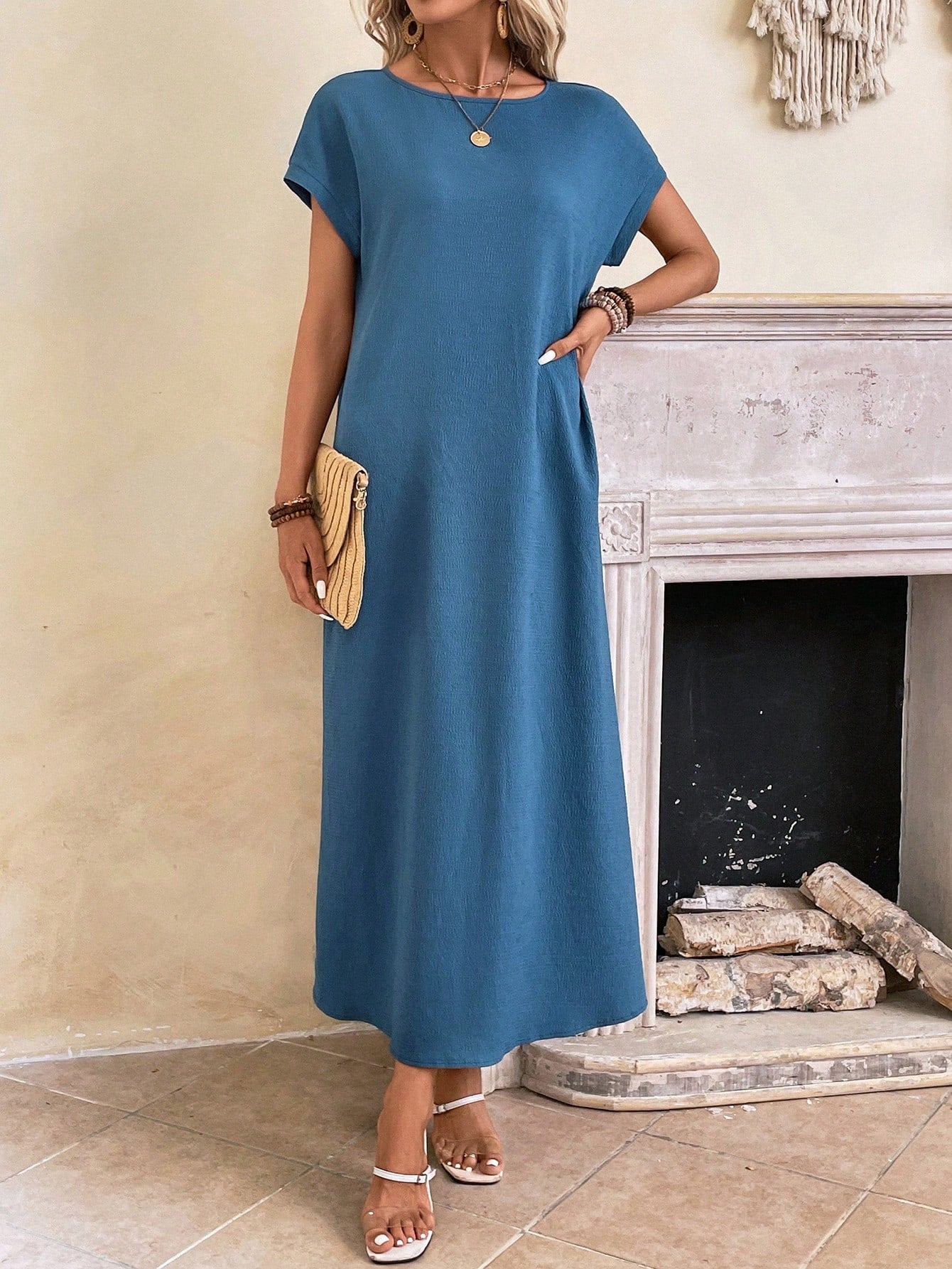 Obscure Blue - A woman wearing a beige fine-knit maxi dress with cap sleeves and a crew neck, styled with gold jewelry and a woven clutch in a minimalist interior.
