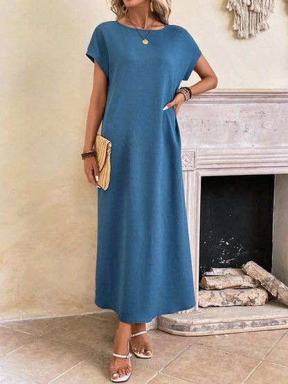 Obscure Blue - A woman wearing a beige fine-knit maxi dress with cap sleeves and a crew neck, styled with gold jewelry and a woven clutch in a minimalist interior.