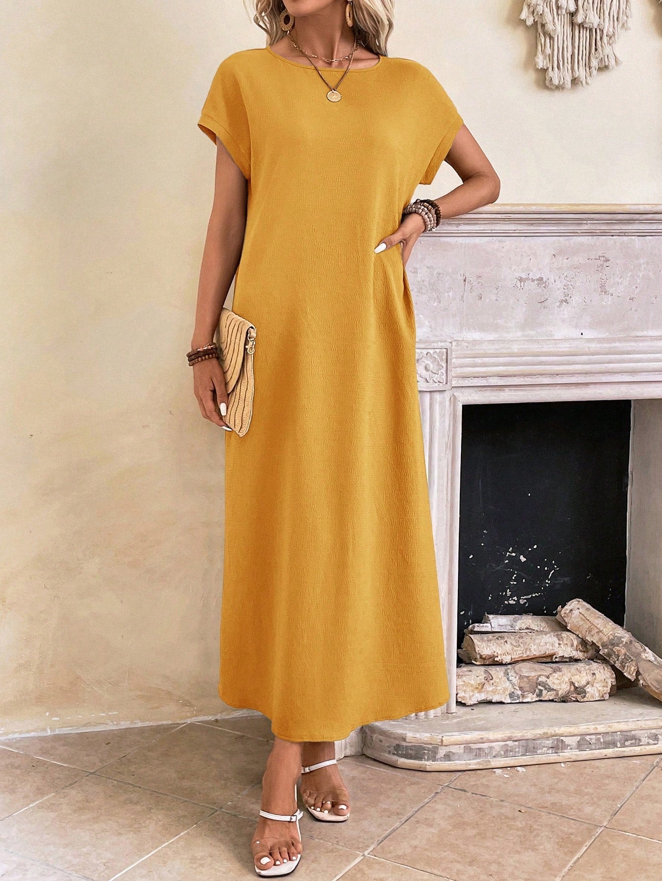 Obscure Blue - A woman wearing a beige fine-knit maxi dress with cap sleeves and a crew neck, styled with gold jewelry and a woven clutch in a minimalist interior.