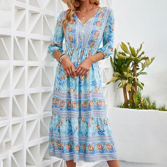 Blue - A woman wearing a light blue midi dress with a botanical floral border print, featuring a V-neckline, three-quarter sleeves, and a tiered skirt.