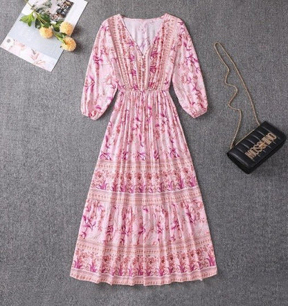 Pink - A woman wearing a light blue midi dress with a botanical floral border print, featuring a V-neckline, three-quarter sleeves, and a tiered skirt.