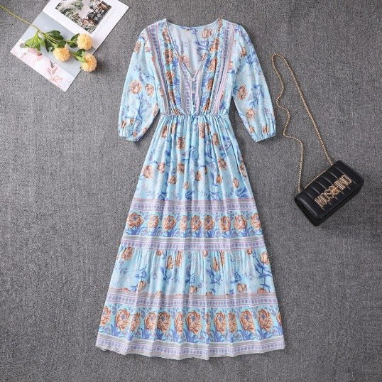Orange - A woman wearing a light blue midi dress with a botanical floral border print, featuring a V-neckline, three-quarter sleeves, and a tiered skirt.