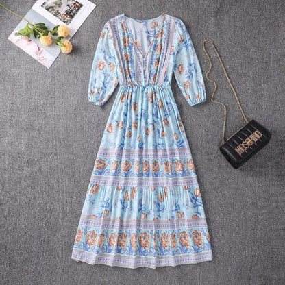 Orange - A woman wearing a light blue midi dress with a botanical floral border print, featuring a V-neckline, three-quarter sleeves, and a tiered skirt.