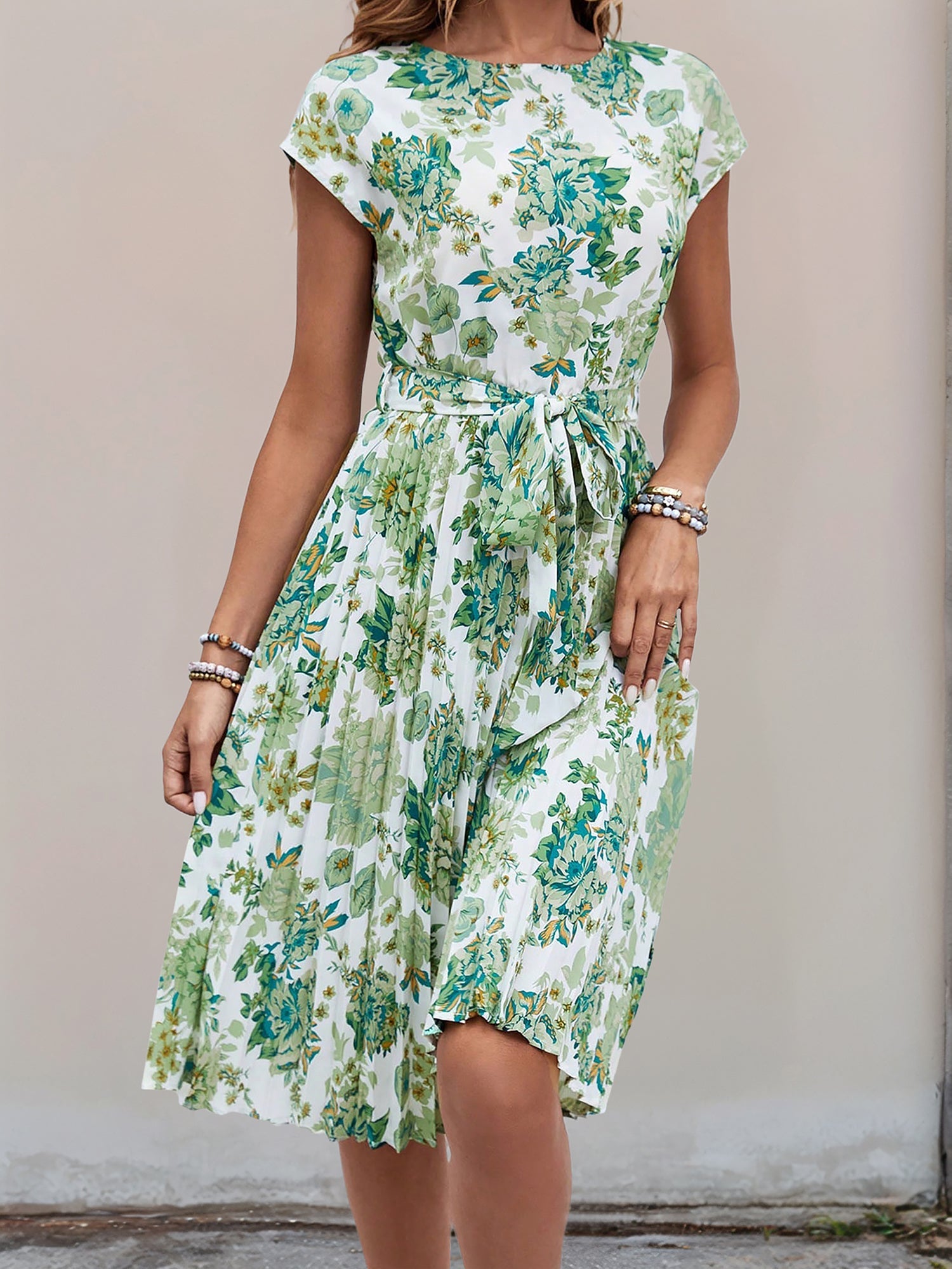 Green - Woman wearing a lightweight floral cap sleeve midi dress with a tie waist and pleated skirt.